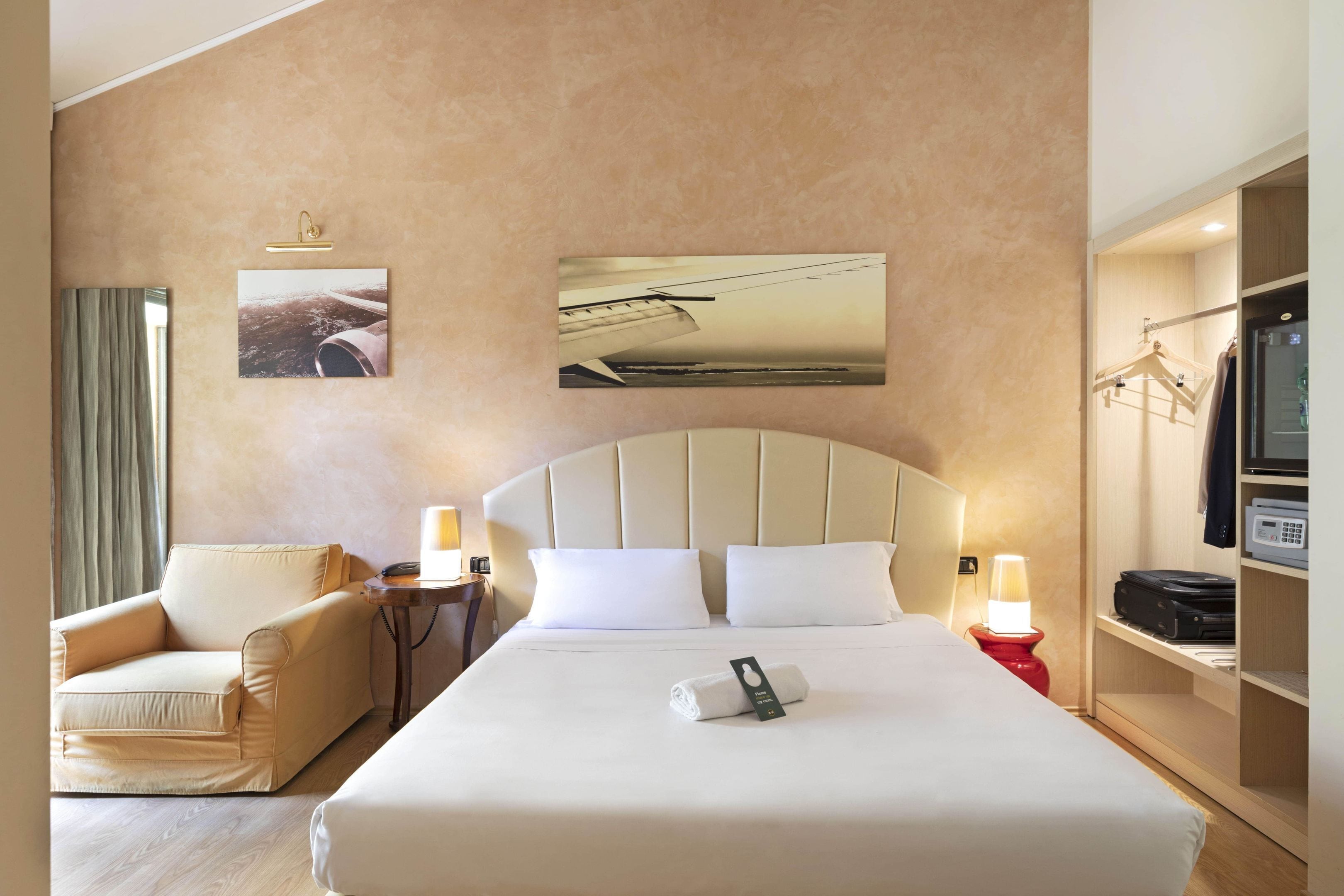 A hotel room with a large bed, a chair, a bedside table with a lamp, and a wardrobe.