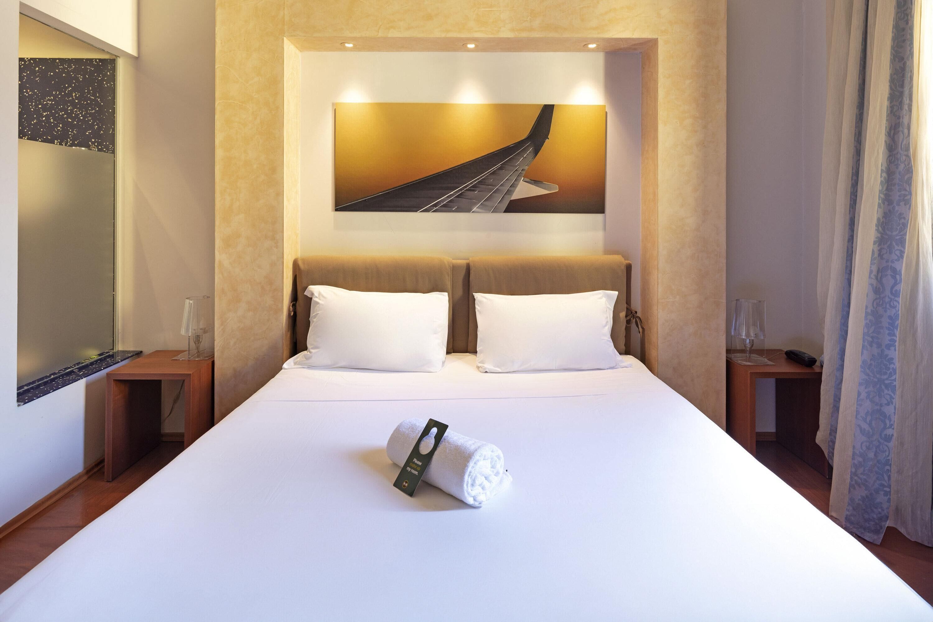 Habitación de hotel con una cama grande, dos almohadas, una toalla, un cuadro de un puente y una mesita de noche con una lámpara.