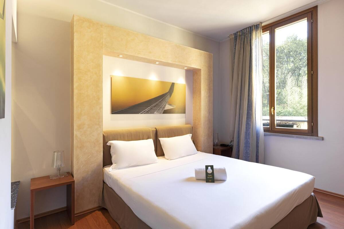 A hotel room with a large bed, a nightstand, a framed picture above the bed, a window with curtains, and a view of trees outside.