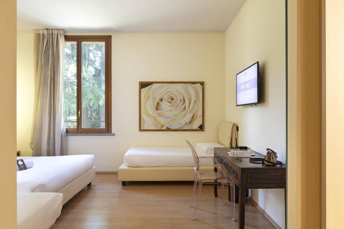 A hotel room with two beds, a desk, a chair, a TV, and a large flower painting on the wall.