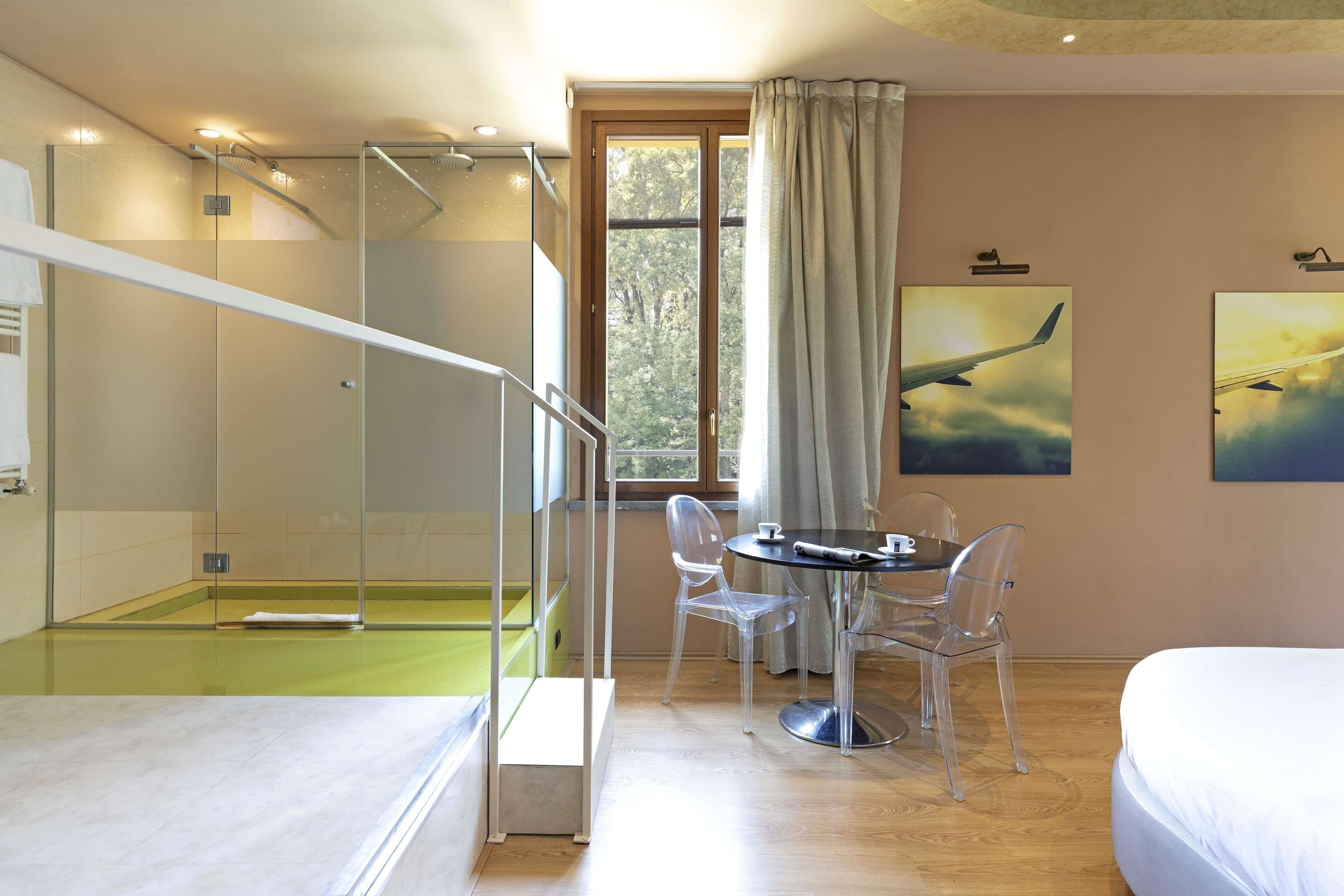 Una habitación de hotel moderna con ducha de vidrio, zona de comedor con mesa y sillas, cama y ventana grande con vista a árboles.