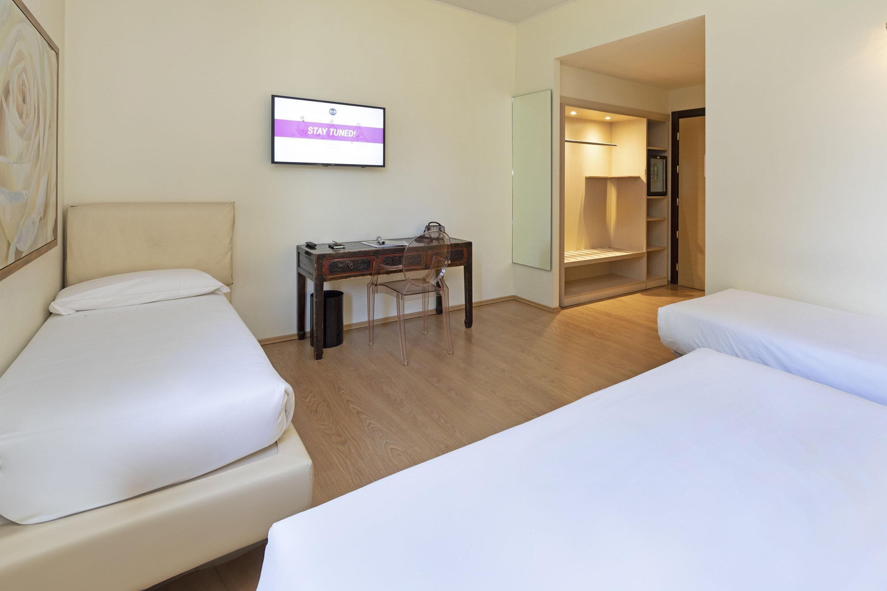 Quarto de hotel com duas camas, uma escrivaninha, uma cadeira, uma televisão e um espelho.