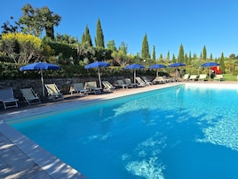 Una piscina con lettini e ombrelloni blu, circondata da alberi e un muro in pietra.