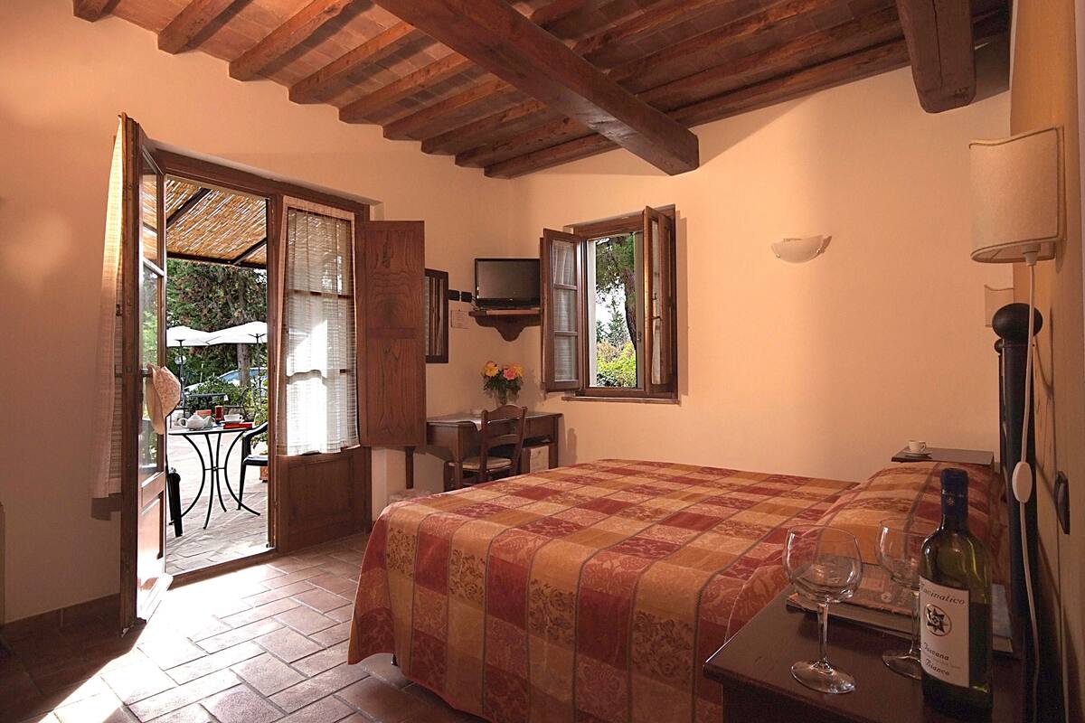 Une chambre à coucher avec un lit, une télévision, une table avec une bouteille de vin et des verres, et une fenêtre donnant sur l’extérieur.