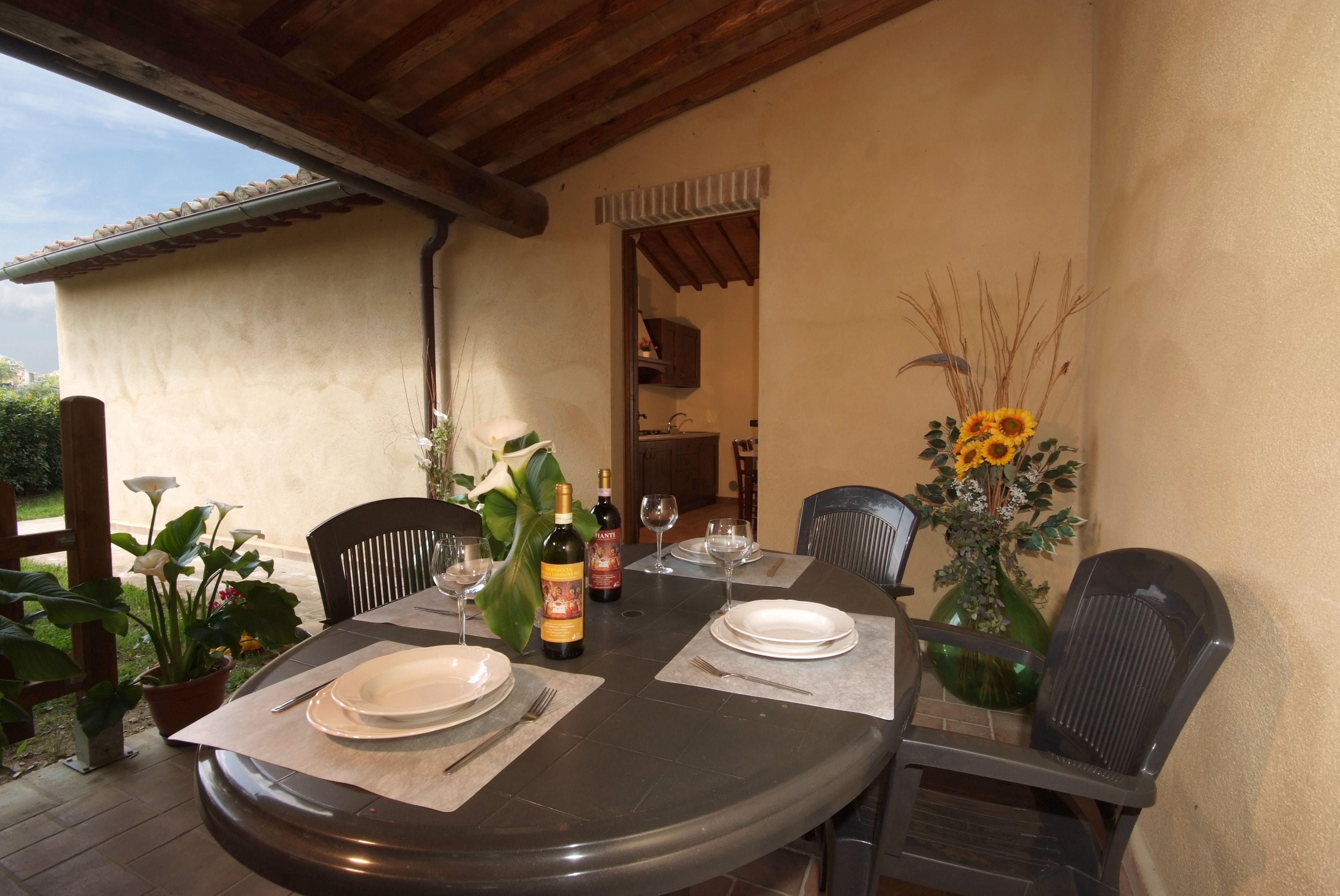 A round dining table set for two with plates, cutlery, and wine glasses. There are chairs around the table, and a potted plant with sunflowers is on the table.