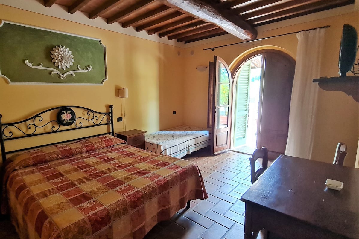 Une chambre à coucher avec un lit, une petite table, un banc, un élément de décoration murale vert et une porte donnant sur l’extérieur.
