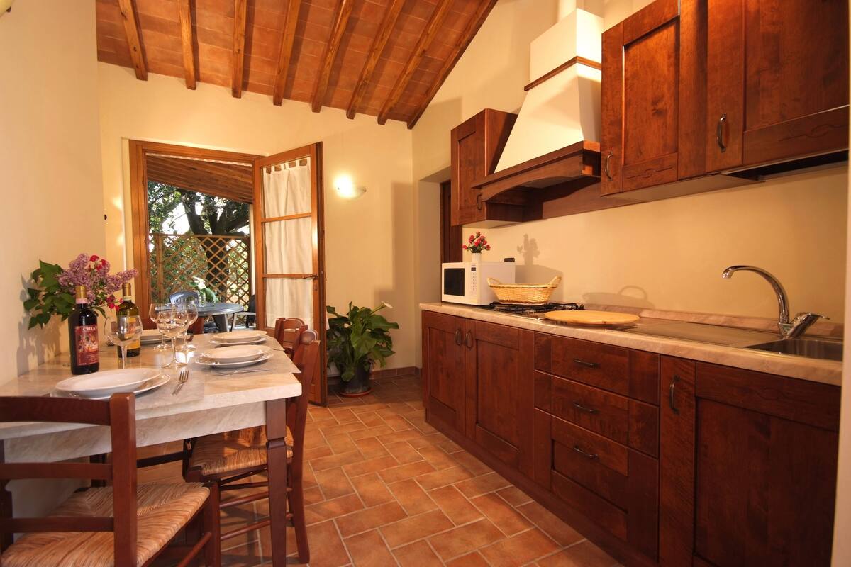 Un coin repas avec une table en bois dressée pour quatre personnes, une cuisine avec des placards en bois et un sol carrelé.