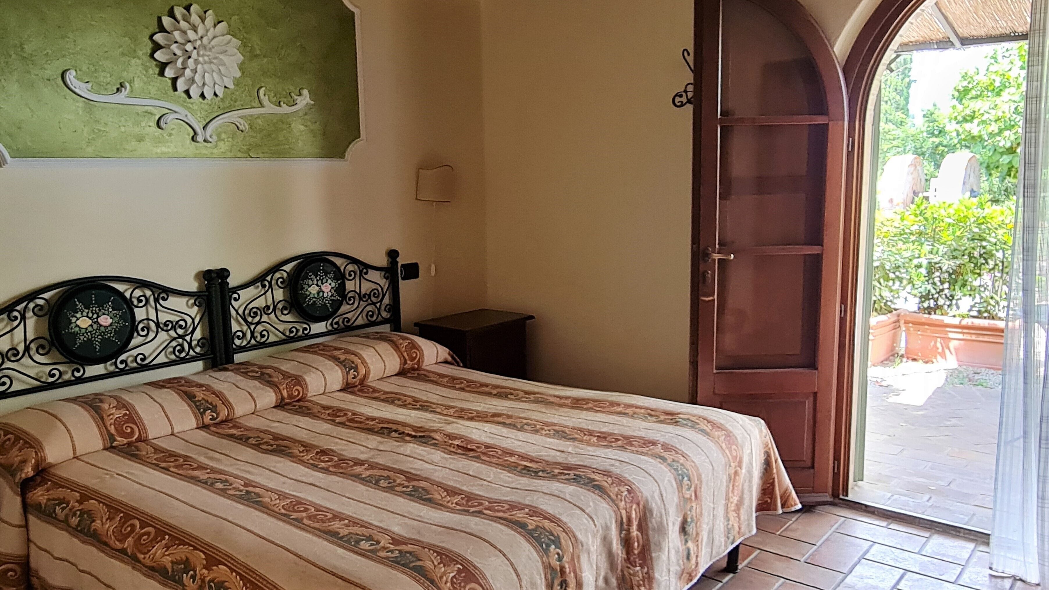 A bedroom with a bed, a nightstand, a green wall with floral decoration, and a wooden door leading to an outdoor area.