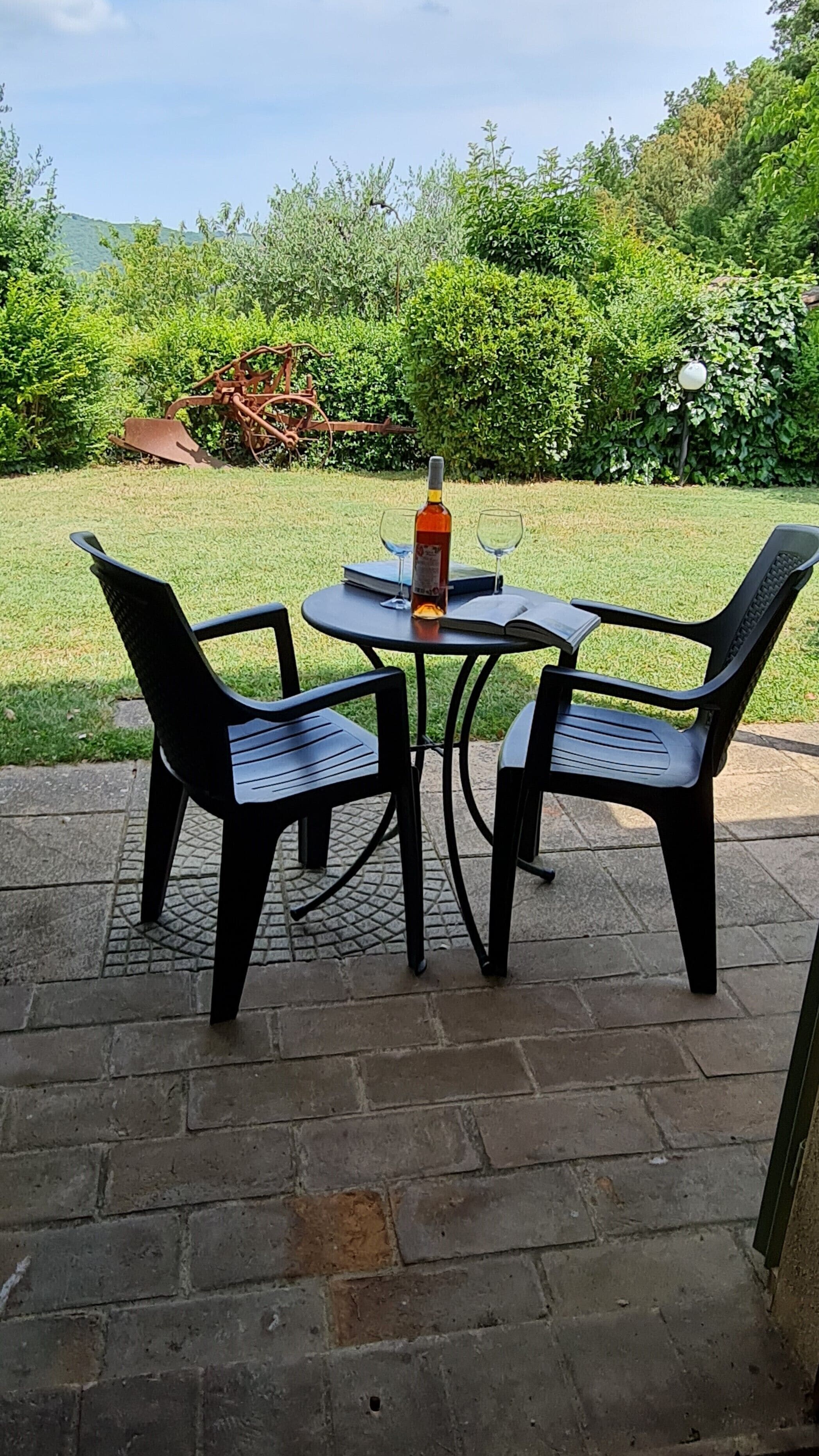 Un patio con dos sillas de plástico negro y una mesa, una botella de vino y dos vasos sobre la mesa.