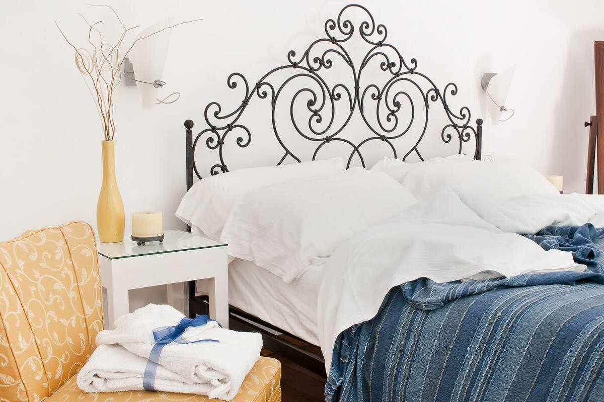Ein ordentlich gemachtes Bett mit weißer und blauer Bettwäsche, ein Nachttisch mit einer Vase und Kerze sowie ein Stuhl mit Handtüchern.