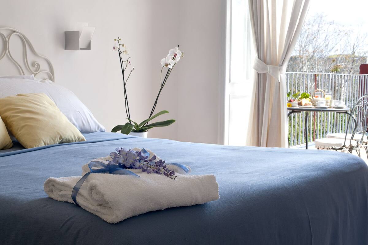 Ein ordentlich bezogenes Bett mit blauer Bettdecke, weißen Kissen und einem Handtuch mit blauer Schleife. Auf dem Nachttisch steht ein Topf mit weißen Blüten.