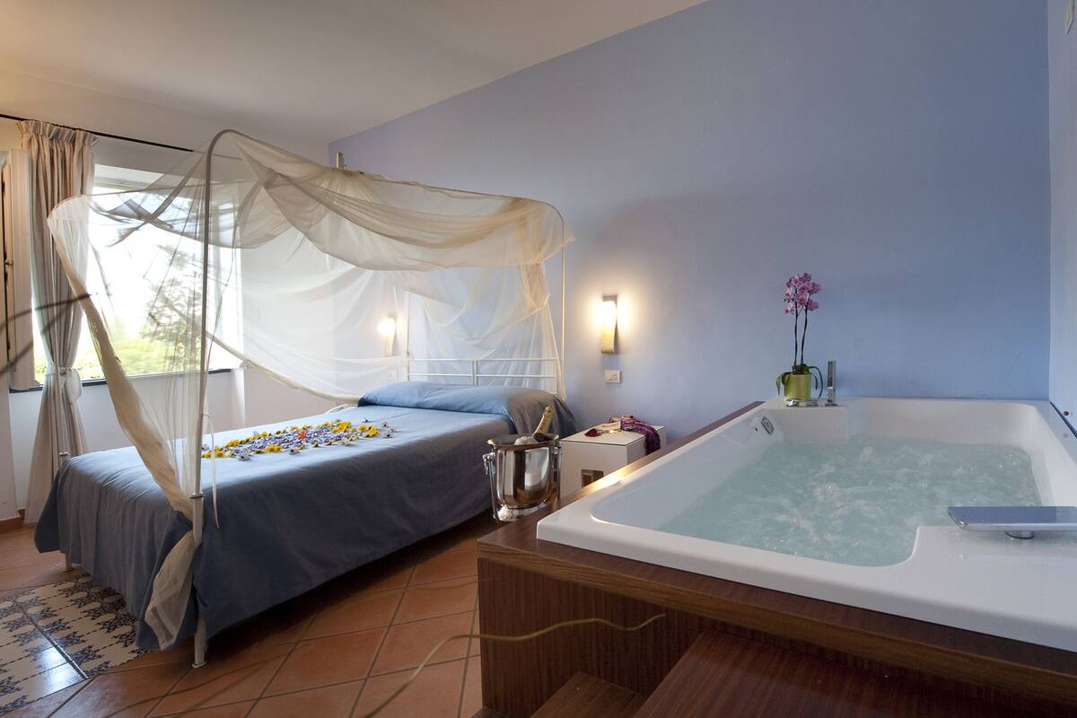 Ein Schlafzimmer mit einem Himmelsbett, einem Nachttisch mit einer Flasche und einer Badewanne mit einer Blumenvase.