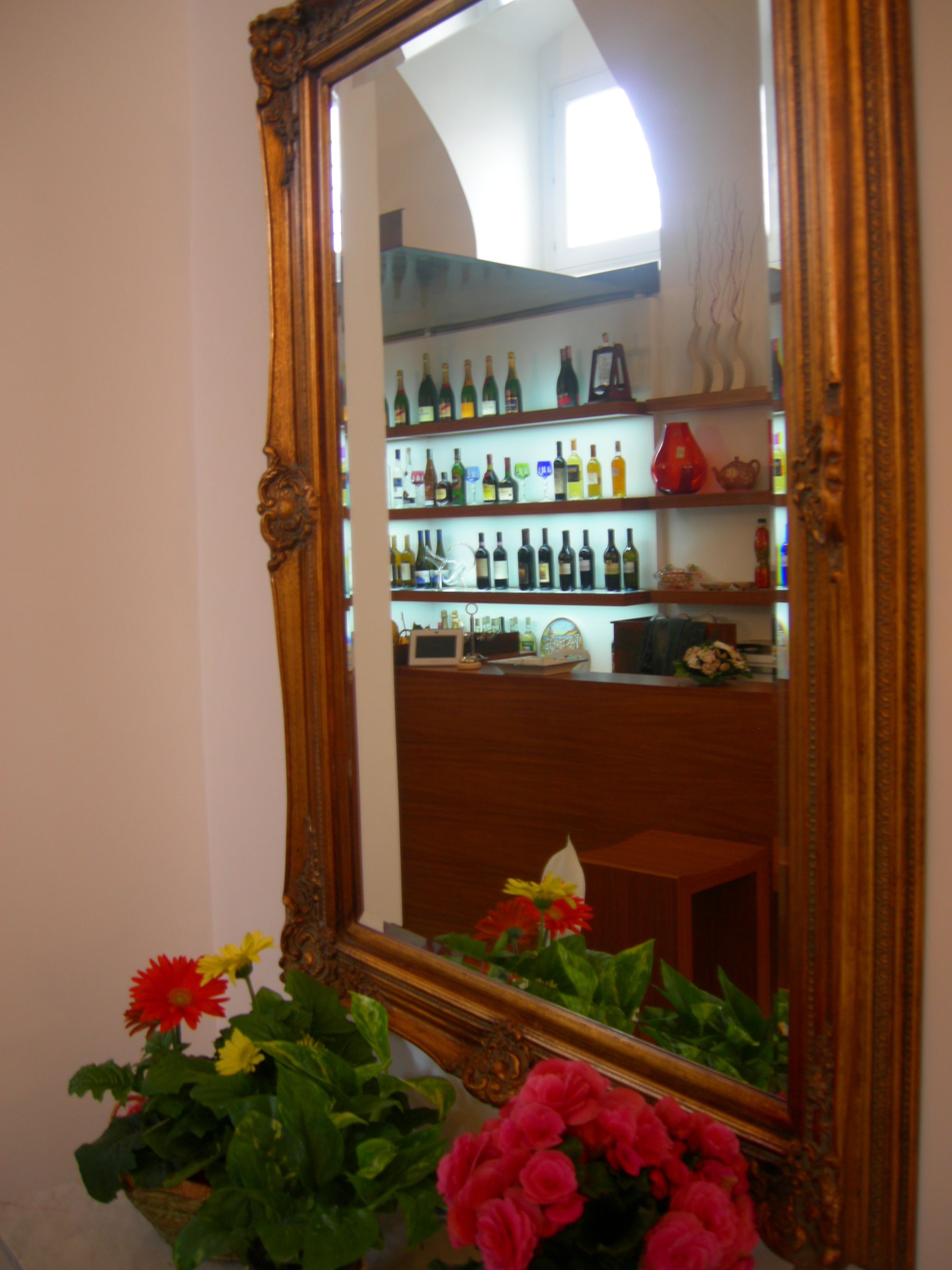 A mirror with a wooden frame reflecting a room with shelves of bottles and a small table.