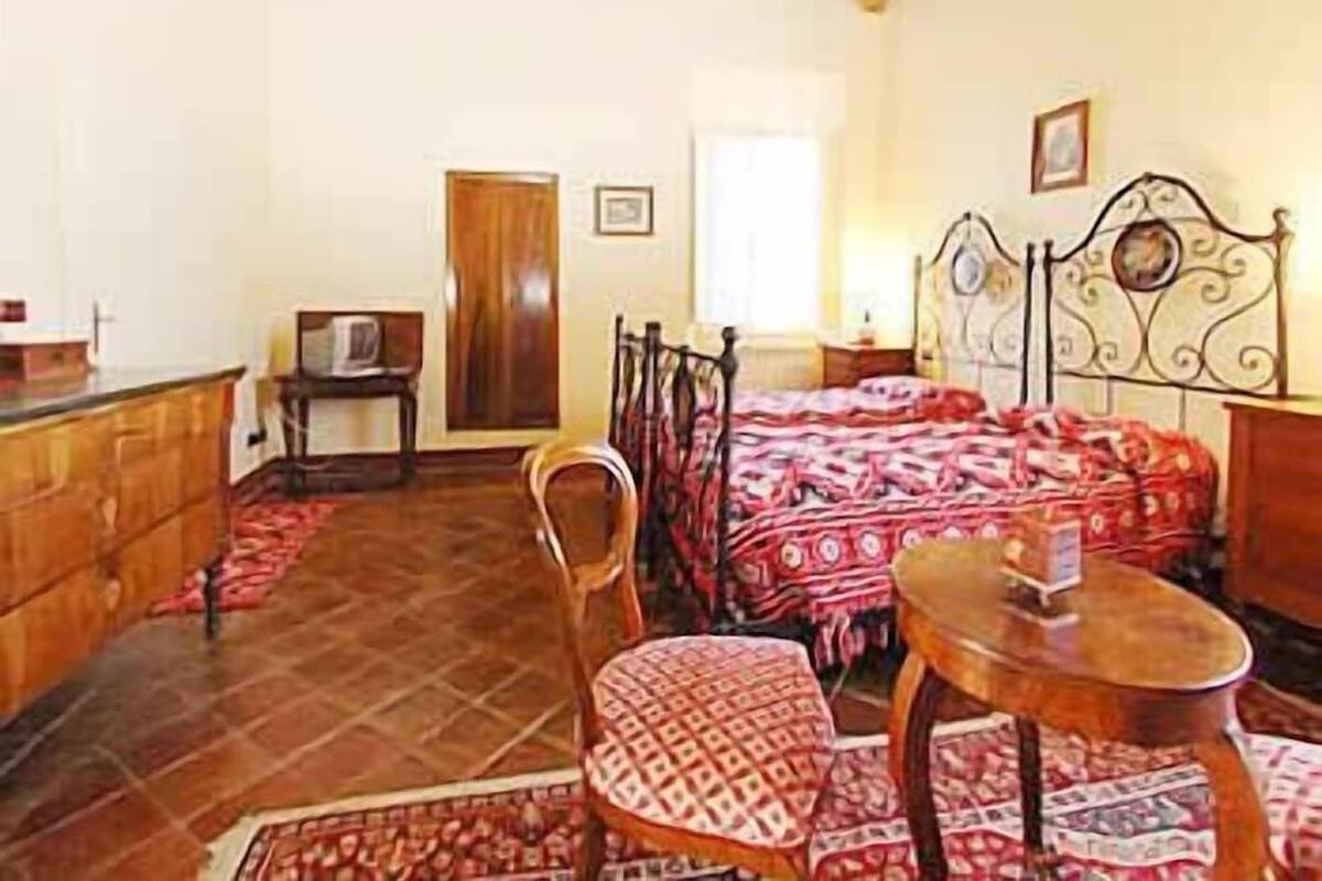 A room with a wooden ceiling, a bed with patterned bedding, a chair, a small table, and a wooden cabinet.