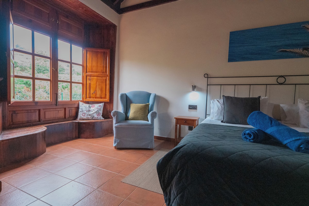 A bedroom with a bed, a chair, a nightstand, a window, and a painting on the wall.