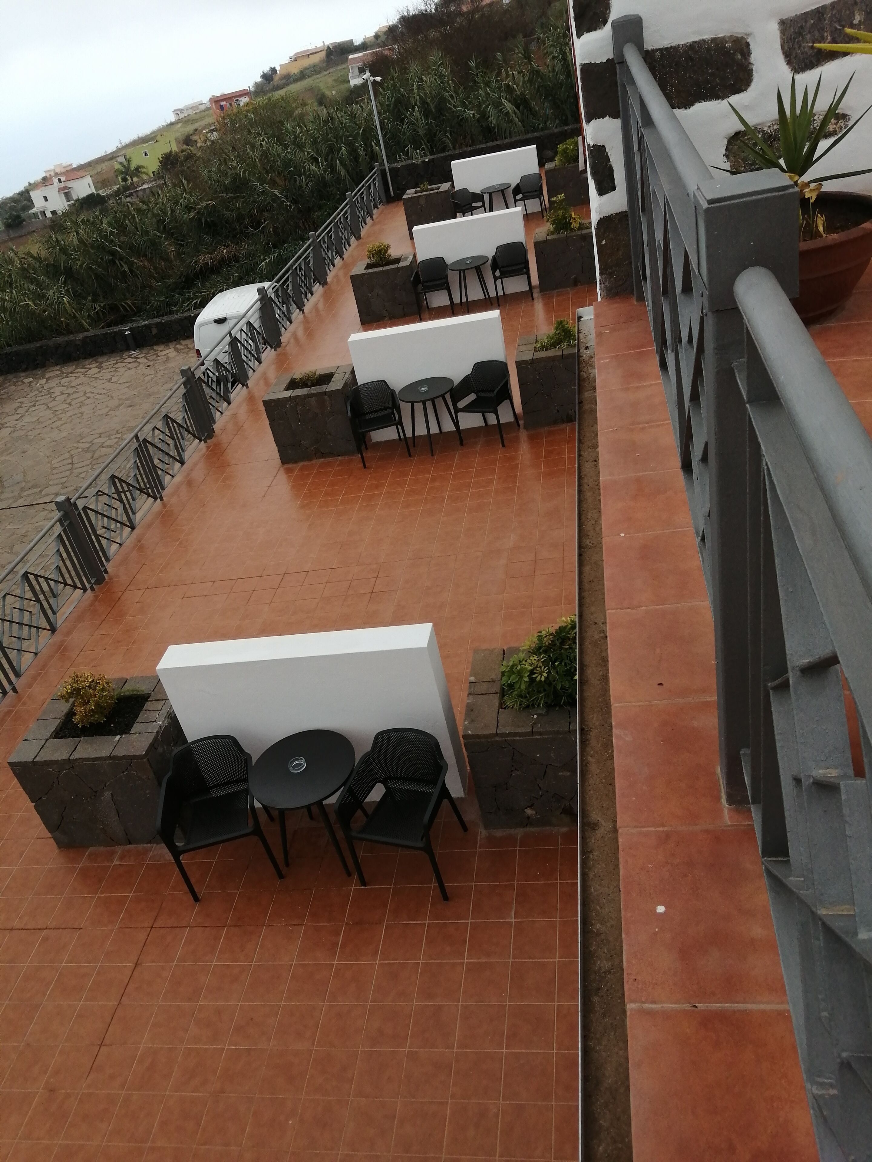 An outdoor patio with multiple tables and chairs, a railing, and a view of the water and greenery in the background.