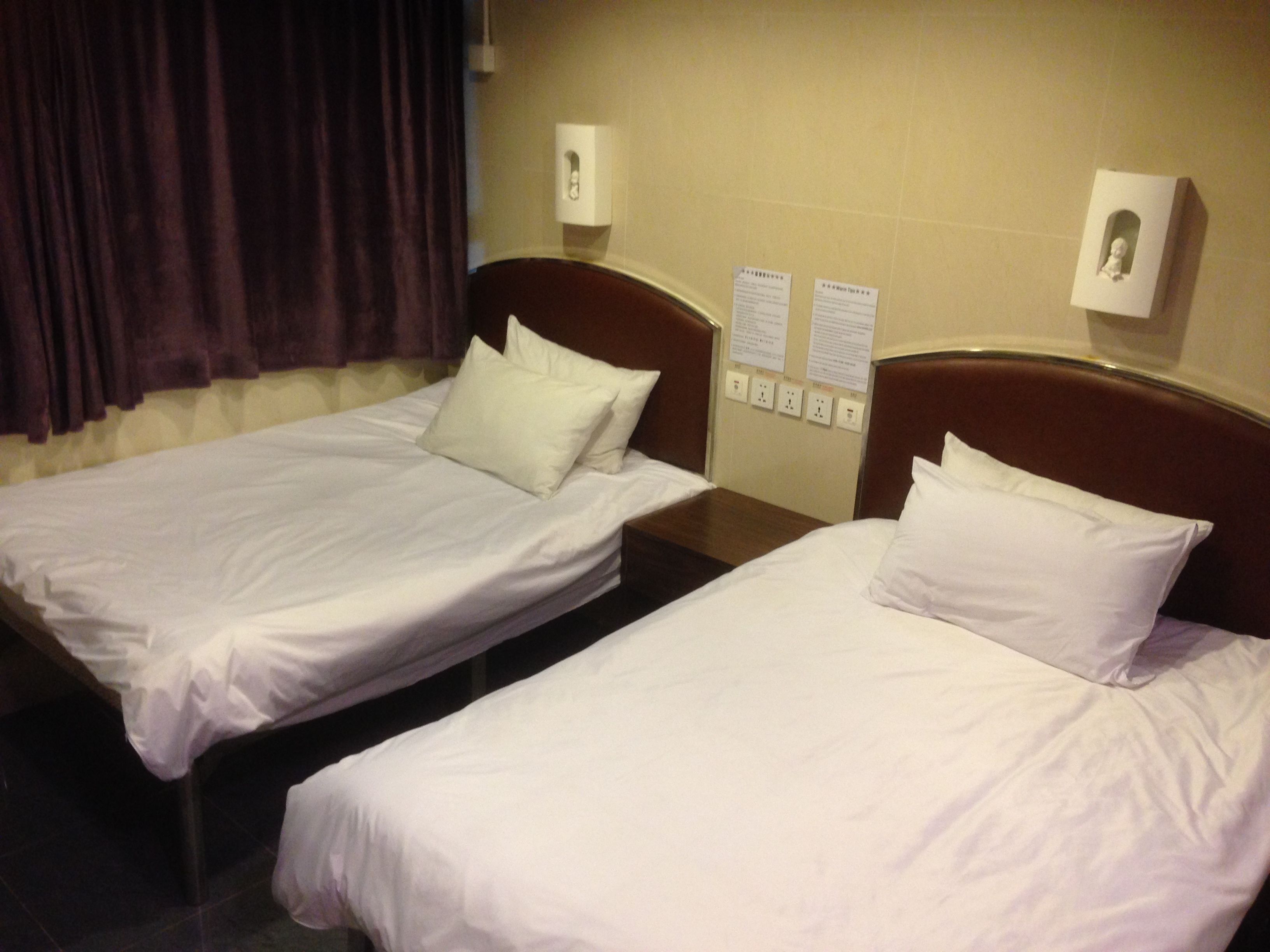 Two single beds with white linens and pillows, a wooden nightstand between them, and a wall-mounted light fixture.
