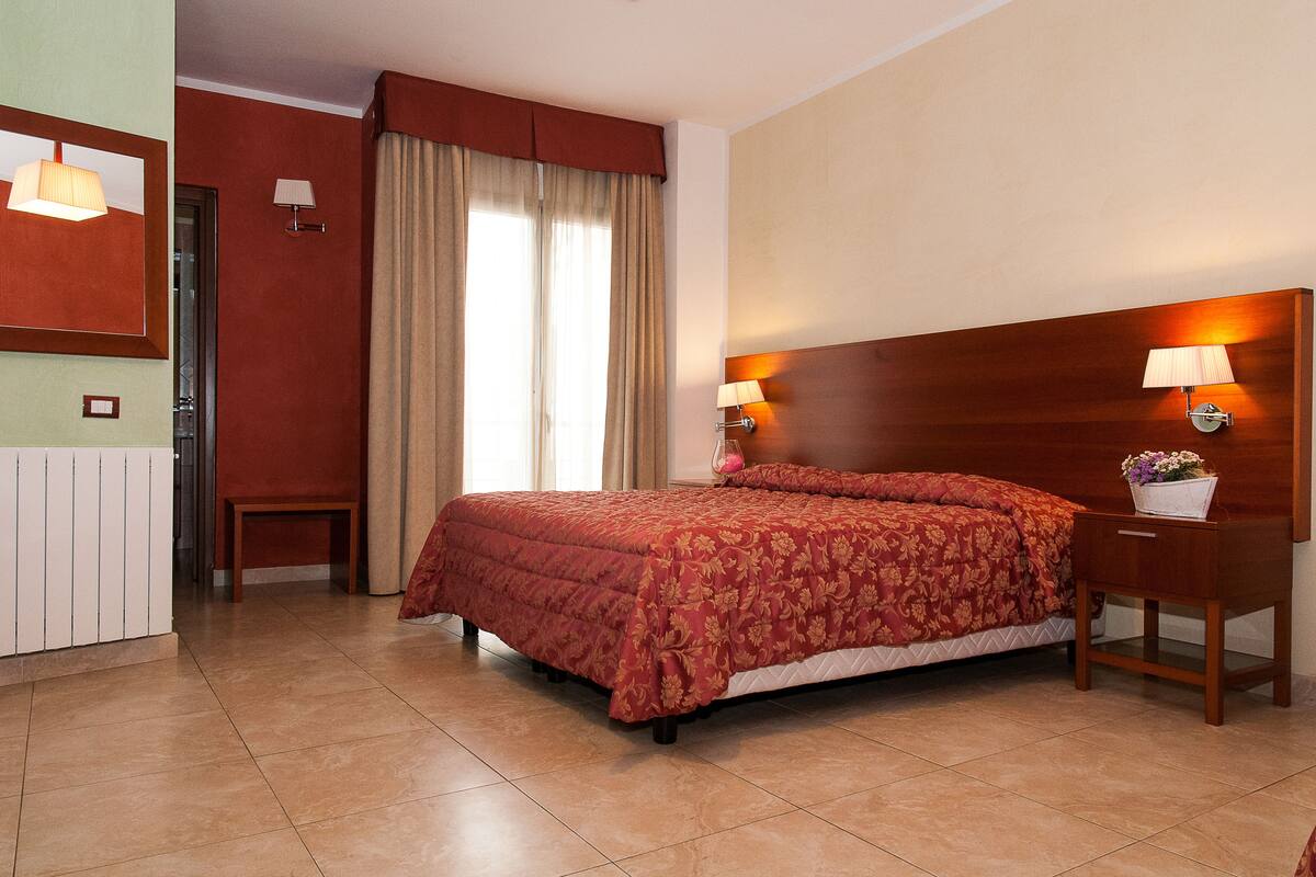 Een hotelkamer met een groot bed, een houten hoofdeinde, twee bedlampen, een klein nachtkastje met een lamp en een spiegel aan de muur.