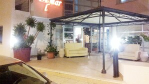 A hotel entrance with a covered waiting area, a neon sign, and potted plants. - Hotel Demar (Olbia)