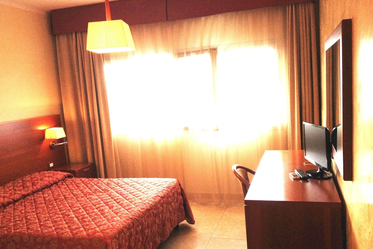 Een hotelkamer met een bed, een bureau met een computer, een lamp en een raam met gordijnen.