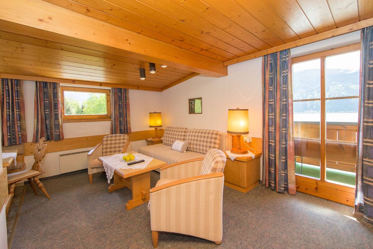 A cozy room with a wooden ceiling, a checkered sofa, a small table with a lamp, and a view of the outdoors through a window.