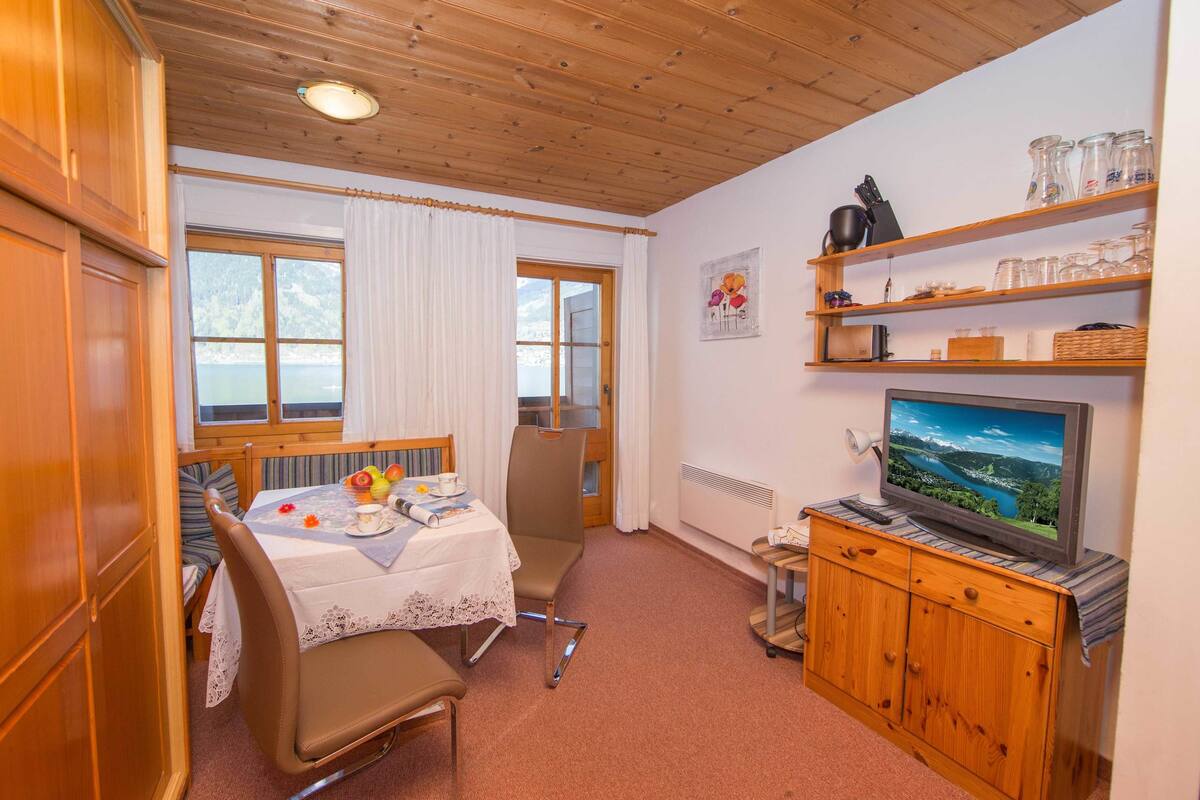 A cozy room with a dining table set for two, a television on a wooden cabinet, and a window with a view of a lake.
