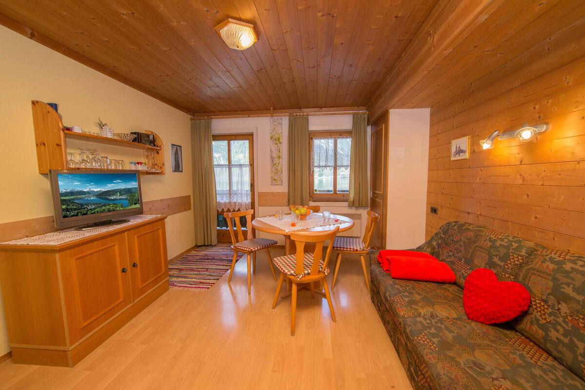 A wooden interior with a dining table, chairs, a sofa, and a television on a wooden cabinet.