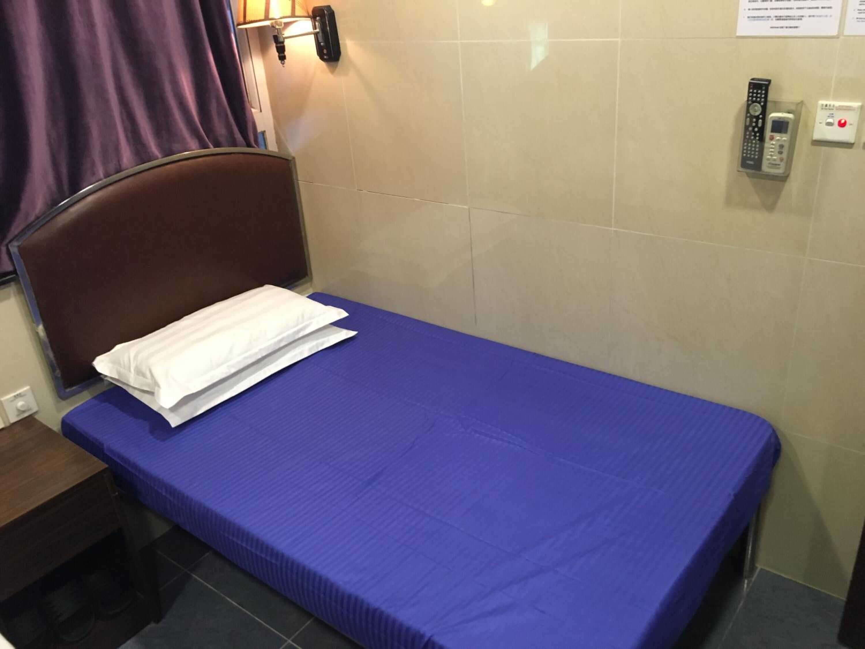 Una cama individual con colchón azul y almohada blanca, cabecera marrón, teléfono montado en la pared y una mesita de noche con lámpara.