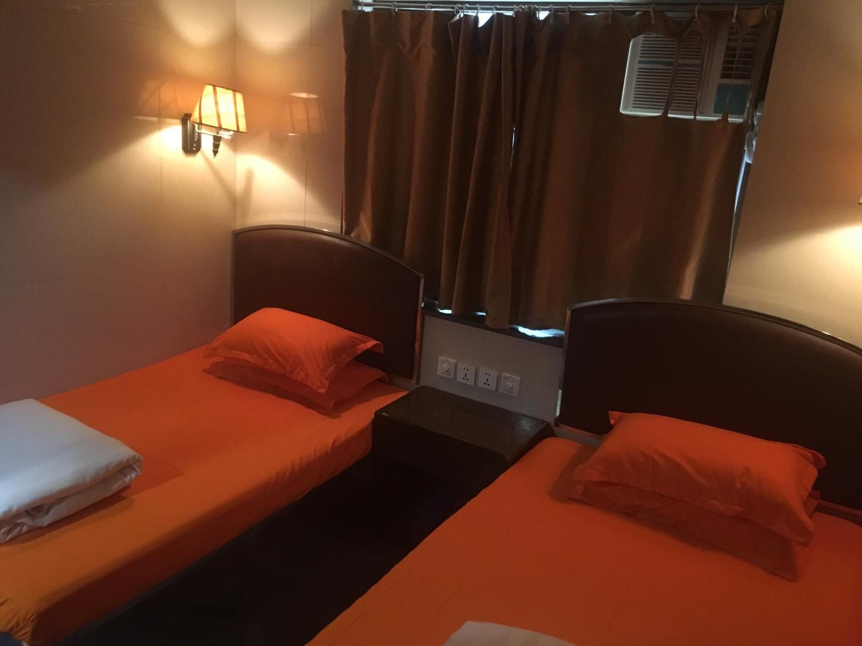 Quarto de hotel com duas camas de solteiro, cada uma com colcha e almofadas laranja combinando. Há luminárias fixadas na parede, uma de cada lado das camas.
