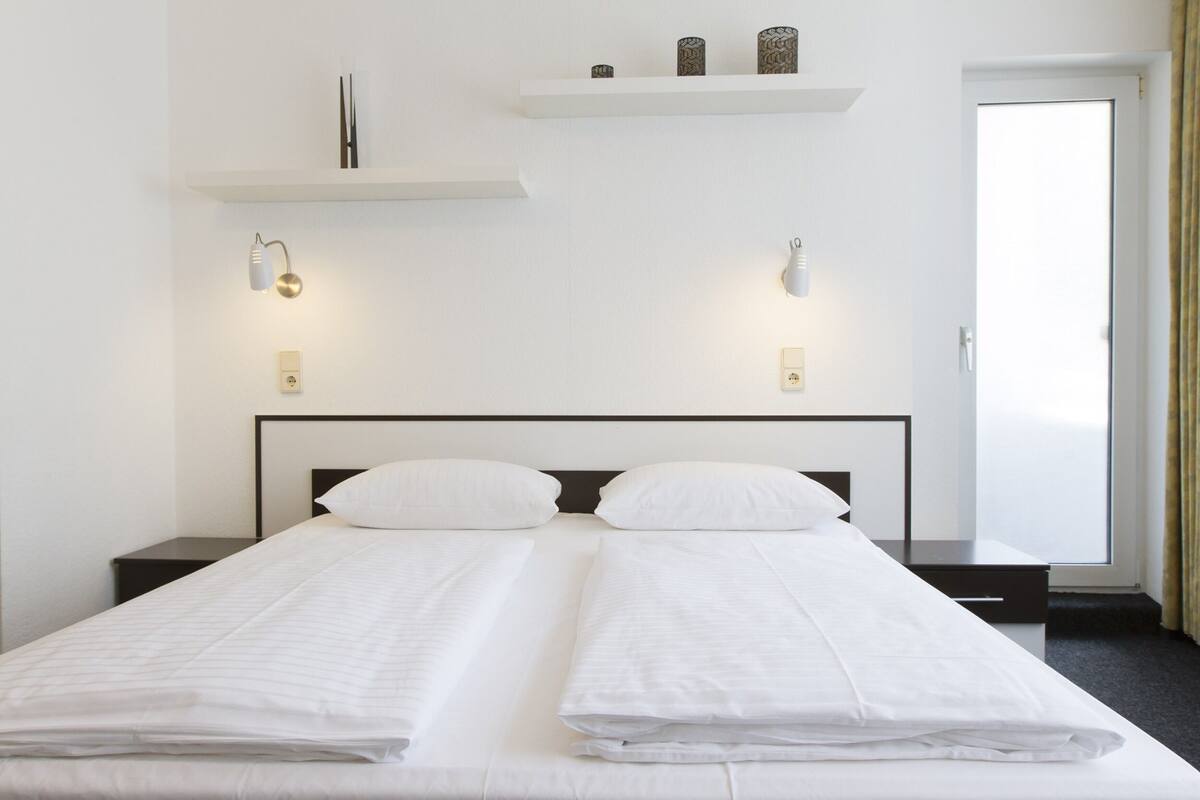 Una cama doble con sábanas blancas, dos mesitas de noche, lámparas de pared y un estante con adornos.