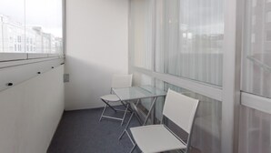 A balcony with a glass table and two white chairs, a white wall, and a view of buildings through large windows. - Aparthotel Vega (Berlin)