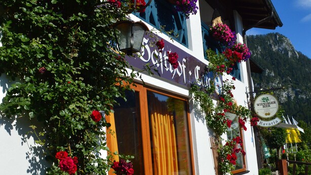 A building with a wooden bench outside, a sign with a logo, and a mountain in the background. - Hotel Gasthaus Café Bavaria (Inzell)