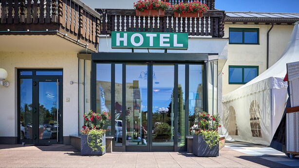 L’entrée d’un hôtel, avec une grande porte vitrée, un balcon orné de jardinières et une enseigne indiquant « HÔTEL ».