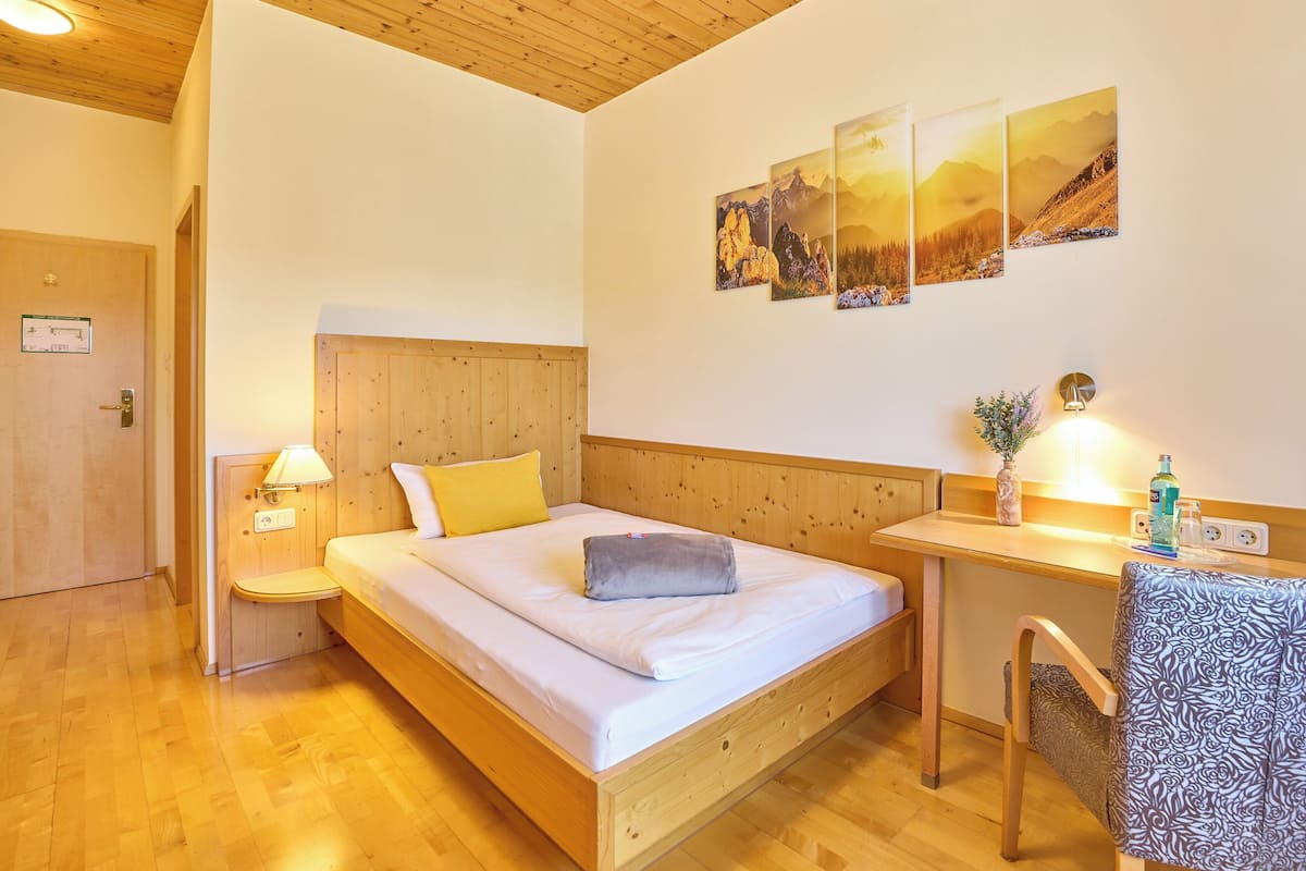 Una cama de madera con cabecera, una mesita de noche con lámpara, y una silla con funda estampada. Hay tres cuadros enmarcados en la pared.