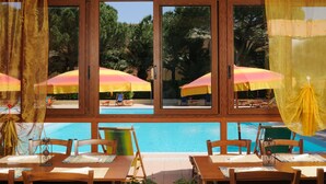 A poolside area with wooden tables and chairs, yellow and pink umbrellas, and a view of the pool and trees. - Hotel Oasis (Alghero)