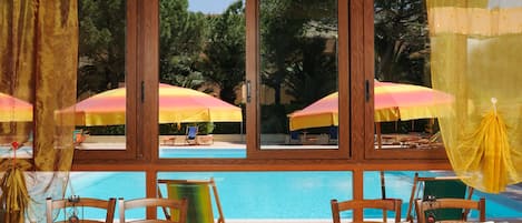 A poolside area with wooden tables and chairs, yellow and pink umbrellas, and a view of the pool and trees.