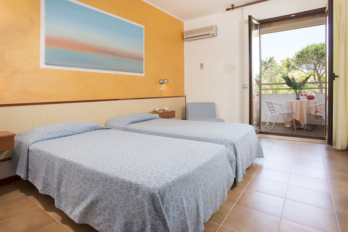 A room with two beds, a painting on the wall, an air conditioning unit, and a balcony with a table and chairs.