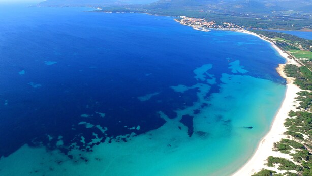 An aerial view of a coastal area with a beach, clear blue waters, and a small town in the background. - Hotel Oasis (Alghero)