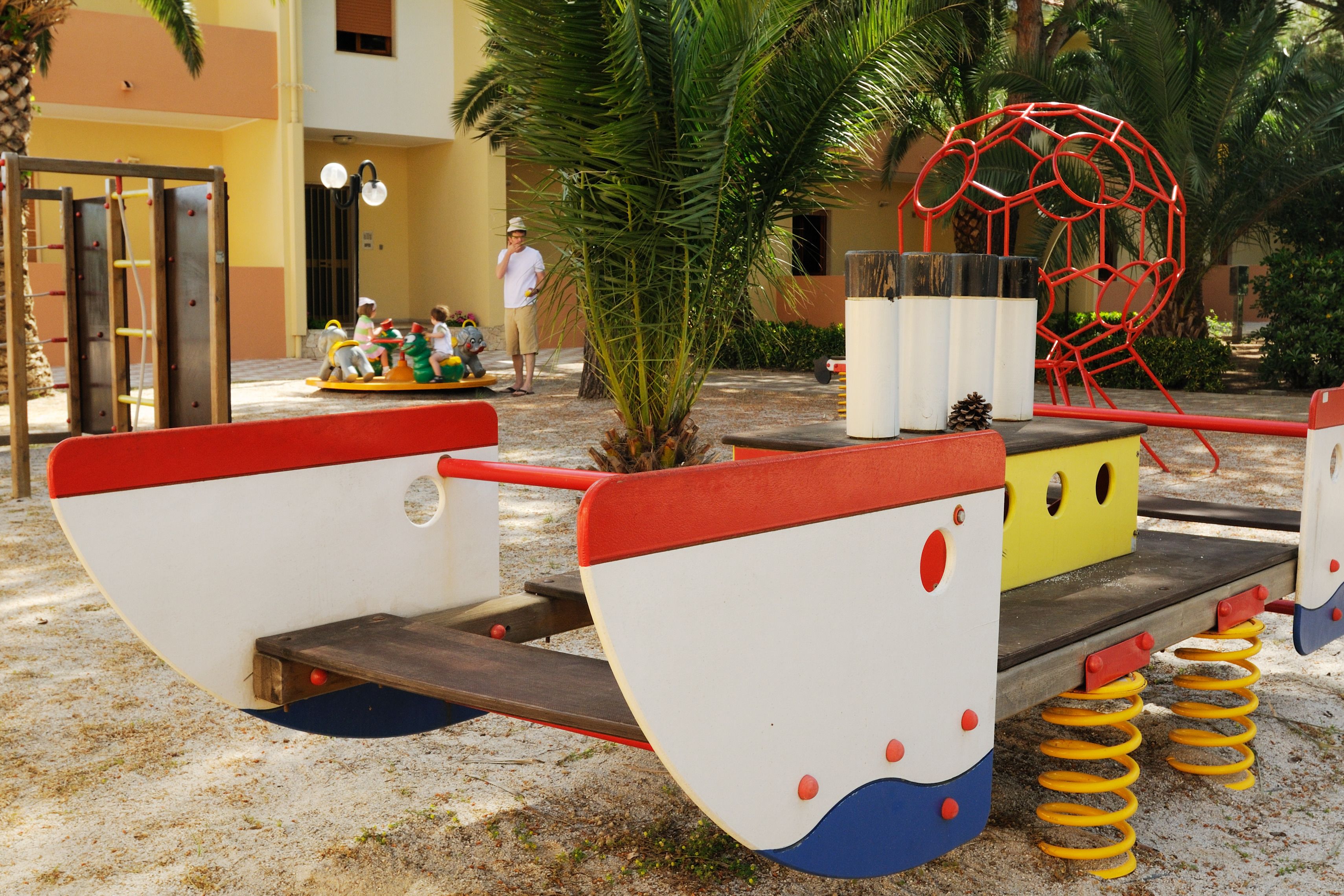 Un parque infantil con un sube y baja, una estructura roja para escalar y un vehículo de juego amarillo y rojo.