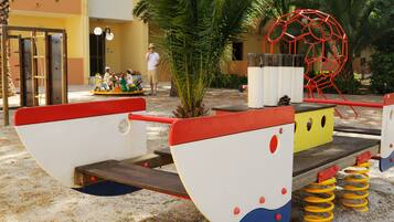 Un parque infantil con un sube y baja, una estructura roja para escalar y un vehículo de juego amarillo y rojo.