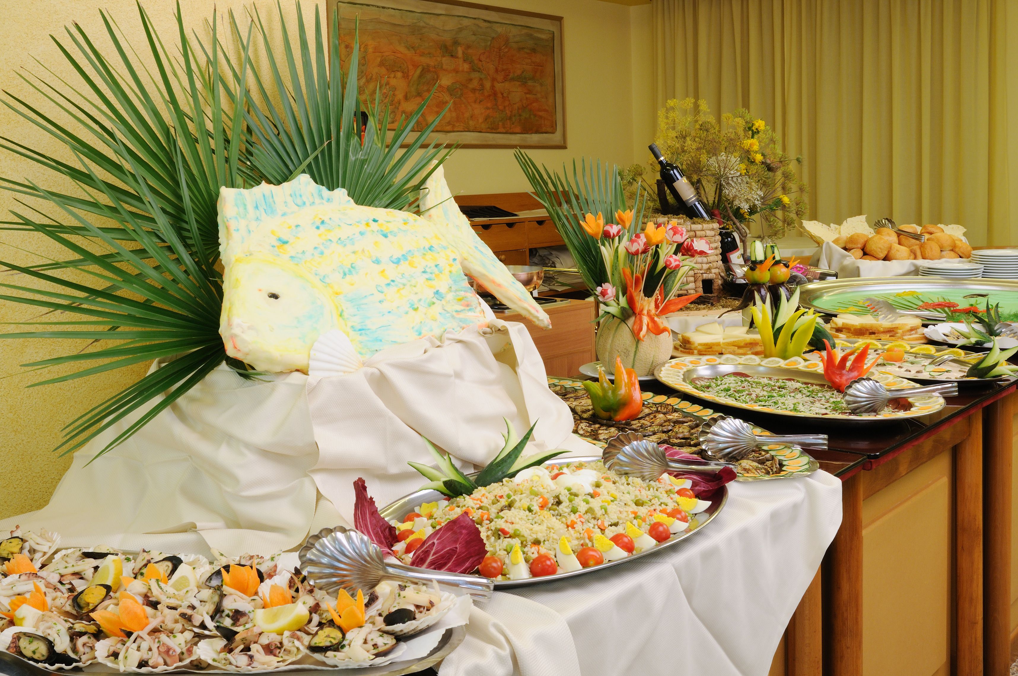 Ein reichhaltiges Buffet mit verschiedenen Speisen, darunter Salate, Früchte und Gebäck, vor einer gelben Wand mit einem Gemälde.