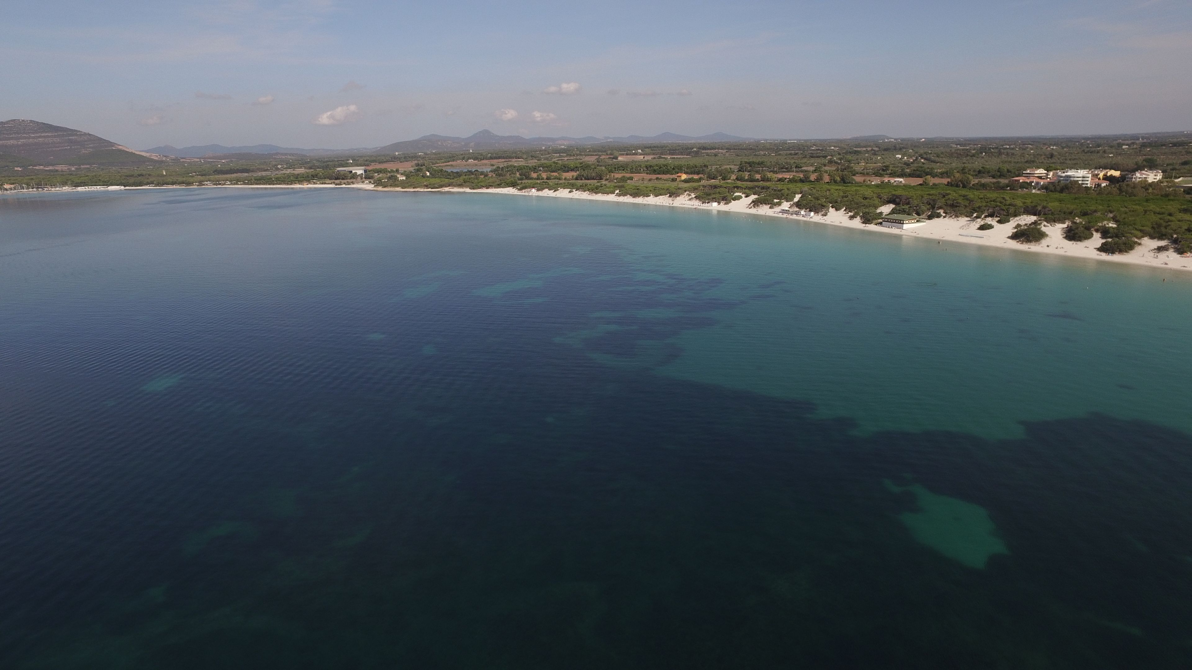 Vista aérea de una zona costera con aguas azules cristalinas, playas de arena y montañas a lo lejos.