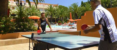 A table tennis court with a net, surrounded by palm trees and a pool area with lounge chairs and umbrellas.