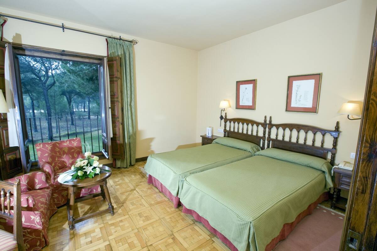 Een hotelkamer met twee bedden, een stoel, een klein tafeltje met bloemen en uitzicht op bomen door een raam.