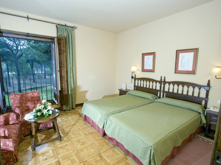 A hotel room with two beds, a chair, a small table with flowers, and a view of trees through a window.