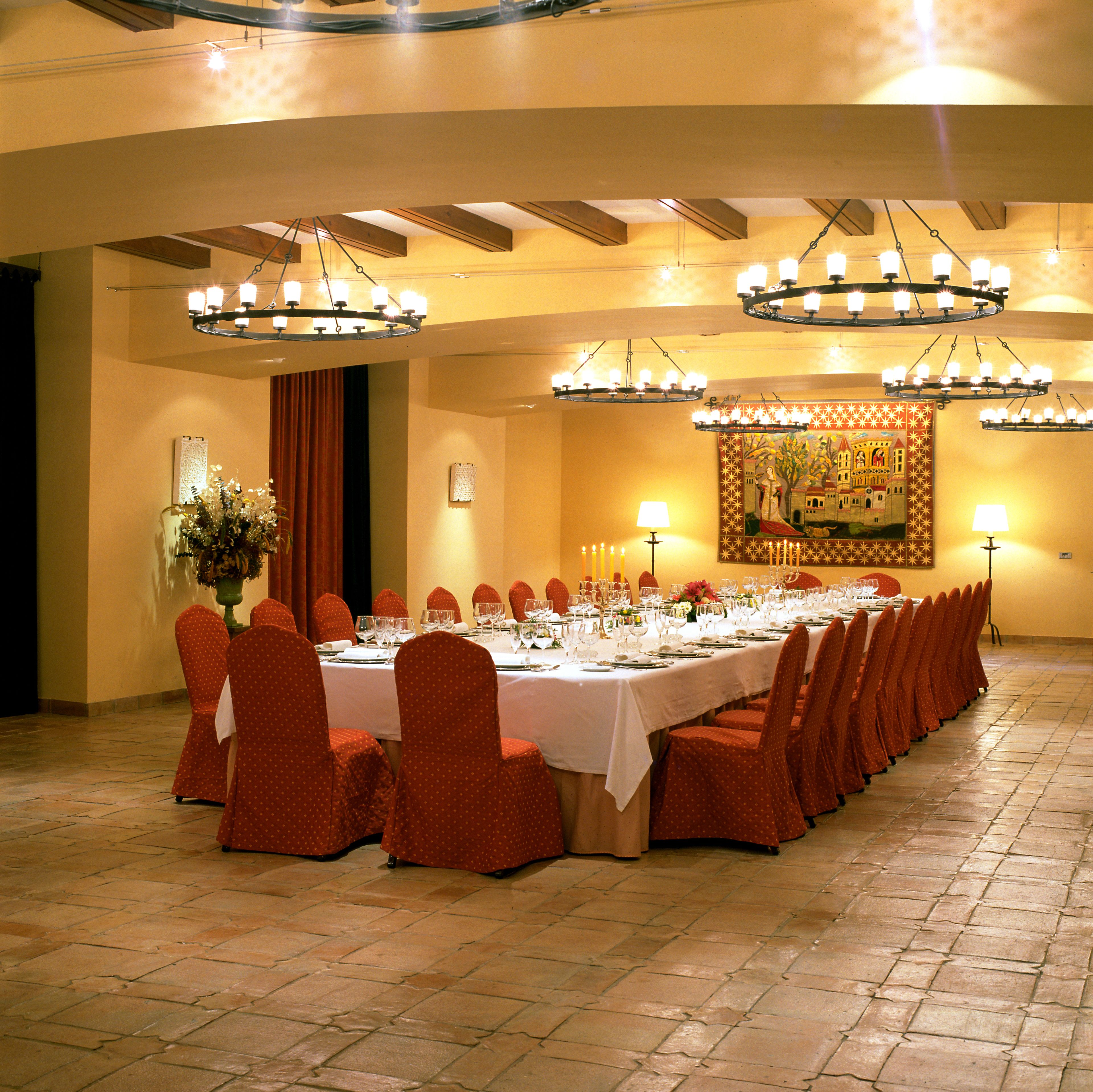 A banquet hall with a long table set for a formal event, surrounded by red chairs, under multiple chandeliers.