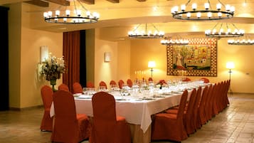 Un salón de banquetes con una larga mesa dispuesta para un evento formal, rodeado de sillas rojas, bajo varios candelabros.