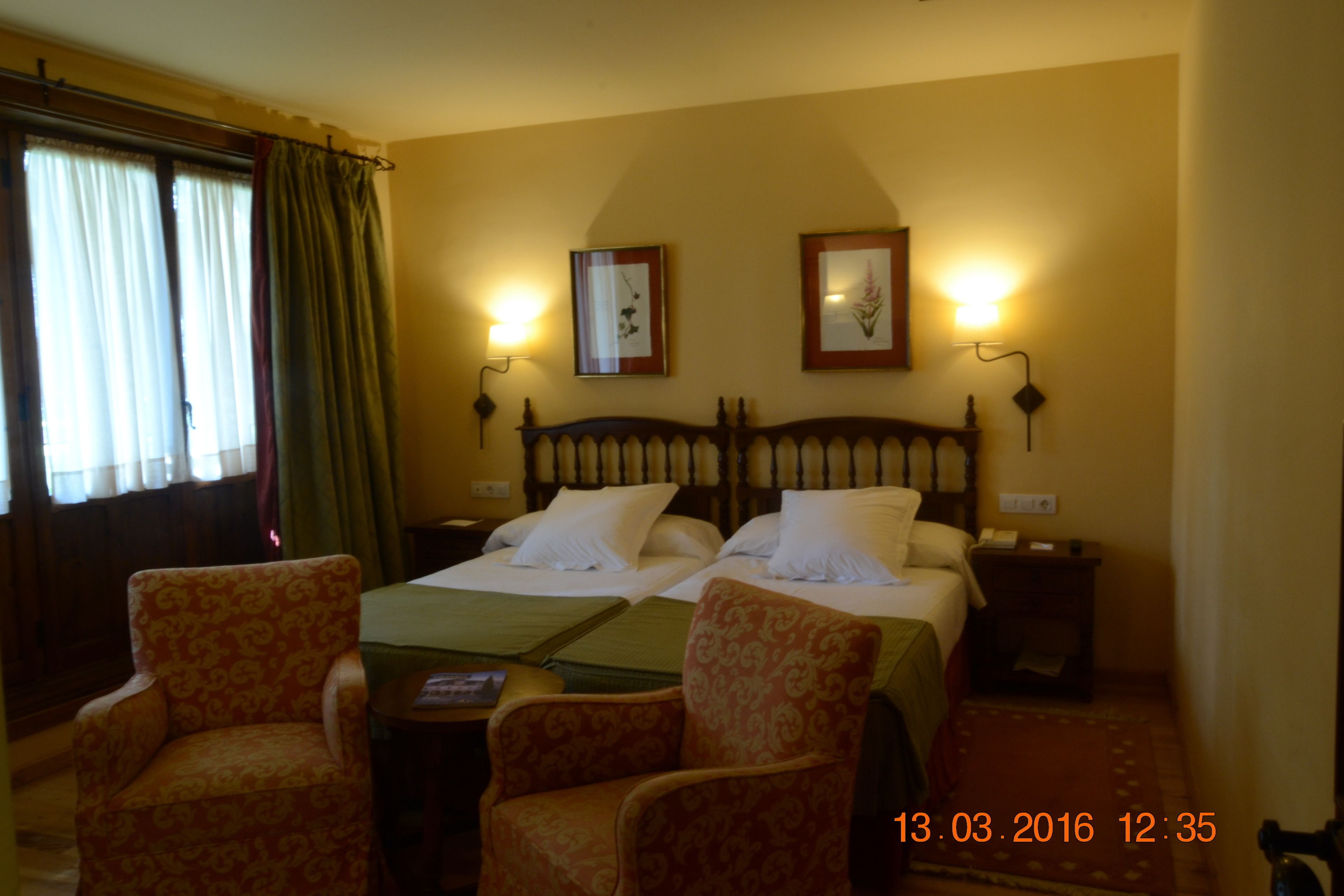 Camera d'albergo con due letti, una testiera in legno, due lampade a parete, un tavolino e due poltrone a motivi decorativi.