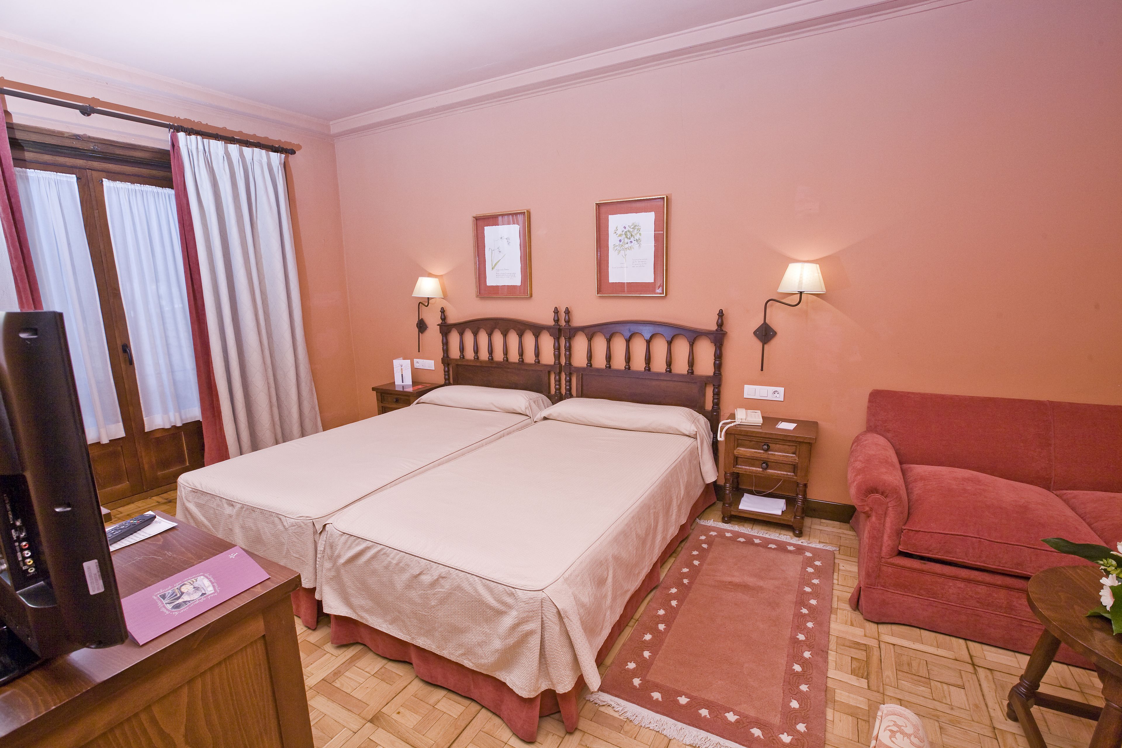 Un lit double avec une tête de lit en bois, deux tables de chevet avec des lampes, un canapé rouge, une petite table ronde avec un vase et un bureau en bois avec une télévision et un dossier rose.