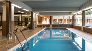 An indoor swimming pool with clear glass walls, wooden pillars, and a view of a building outside.